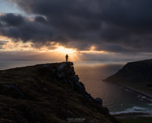 Female hiker watches midnight sun over sea from Nonstind mountain peak, Vestvågøy, Lofoten Islands, Norway